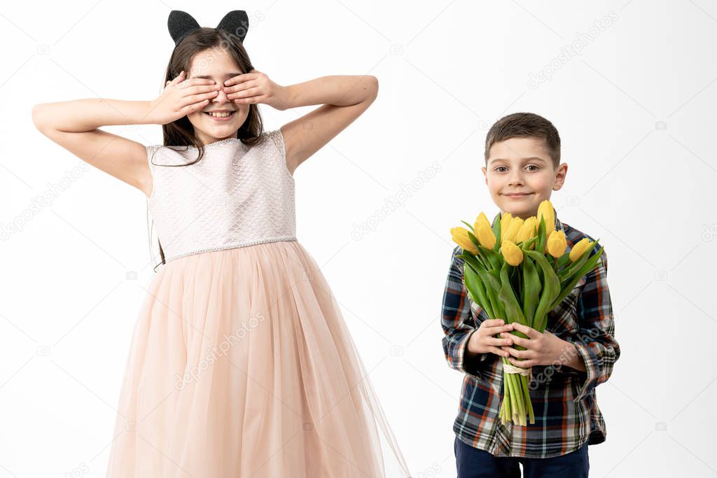 Beautiful happy smiling brunette girl in beige dress covered her eyes with her hands and waiting for present, her little brothes holds bouquet of yellow tulips and looking at the camera isolated over