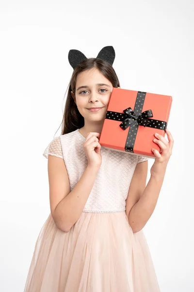Cheerful brunette girl in beige dress holds a gift box in hands near her face and looking at the camera, isolated over white background — Stockfoto