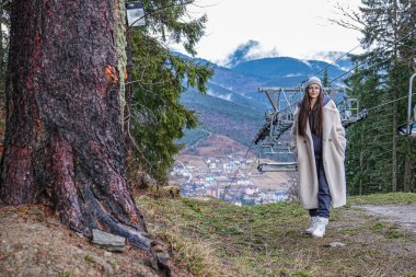 Attractive brunette female in glasses, girl with loose hair dressed in beige coat stands on the top of the hill near the sky-lift and big tree and looking at the camera, amazing view of the mountains