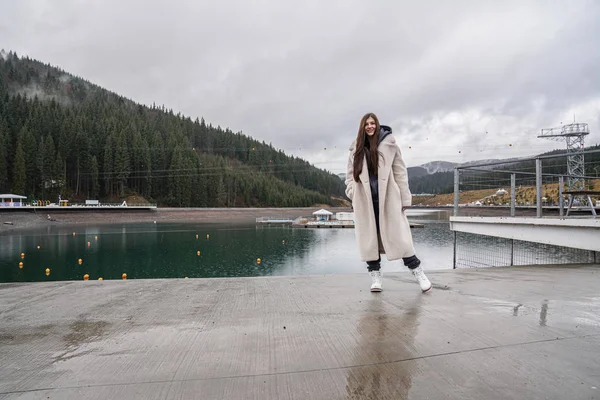 Atraktivní mladá brunetka dívka s volnými vlasy oblečená v béžovém kabátě stojí v blízkosti horského jezera a dívá se na kameru, les v mlze na pozadí — Stock fotografie