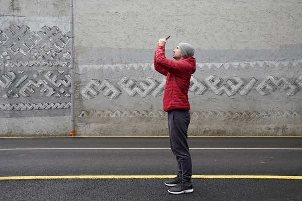 Jovem atraente em casaco vermelho e chapéu cinza fazendo selfie com seu telefone enquanto estava em pé entre a rua, parede de concreto no fundo — Fotografia de Stock
