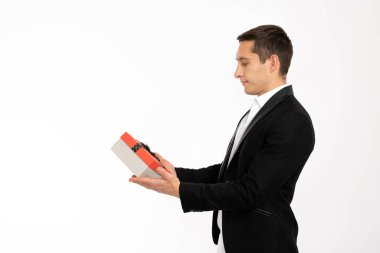 Side view portrait of the attractive confident man in black blazer that holds gift box in hands and looking at it isolated over white background. St. Valentienes day, New Year, Womans day, Christmas