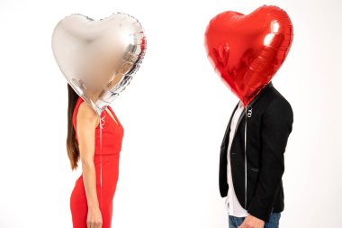Elegant couple stands against each other, covering their faces with silver and red air balloons, St. Valentines day concept, isolated over white background