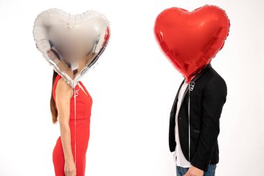 Elegant couple stands against each other, covering their faces with silver and red air balloons, St. Valentines day concept, isolated over white background