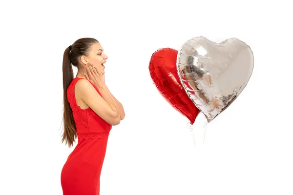 Feliz hermosa chica morena sorprendida en vestido rojo sostiene sus manos sobre sus mejillas, mientras mira dos globos de aire corazones aislados sobre fondo blanco — Foto de Stock