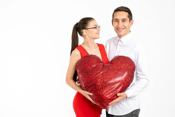 Feliz sonrisa elegante pareja sosteniendo gran corazón leído en las manos, atractiva chica morena en vestido rojo y gafas mirando a su novio aislado sobre fondo blanco con copyspace. Día de San Valentín — Foto de Stock