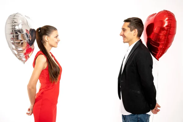 Retrato de pareja romántica encantadora sosteniendo globos de helio en las manos a sus espaldas, los jóvenes elegantes se felicitan mutuamente con el día de San Valentín, disfrutando de tiempo juntos aislados en — Foto de Stock