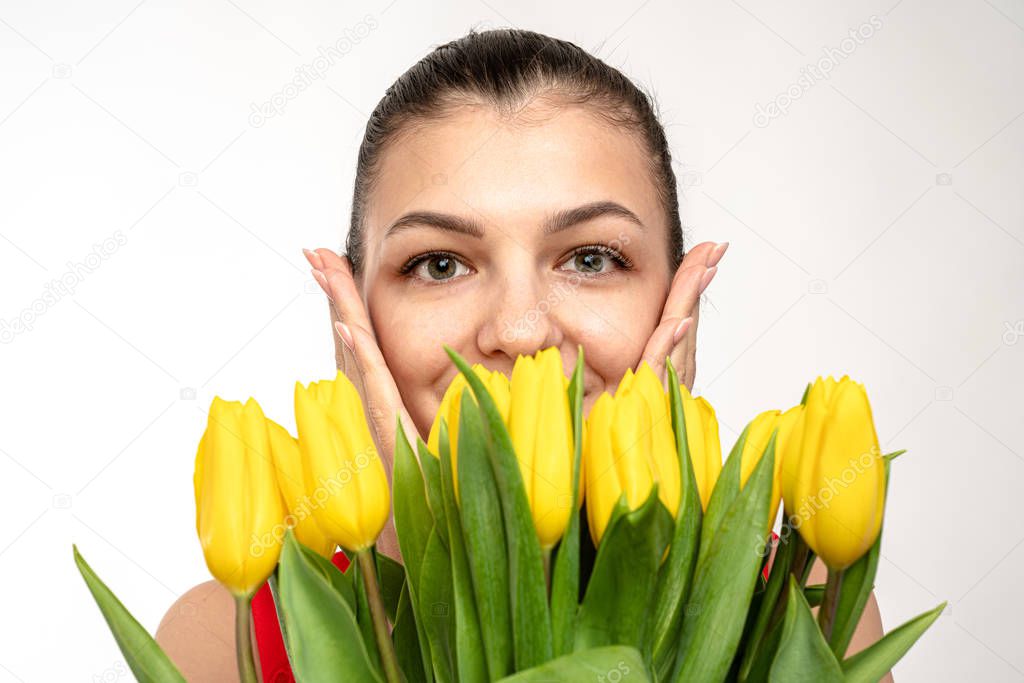 Close up portrait of the brunette young girl with natural healthy skin that holds her hand on her cheeks and looking at the camera, yellow tulips covered half of females face, isolated over white