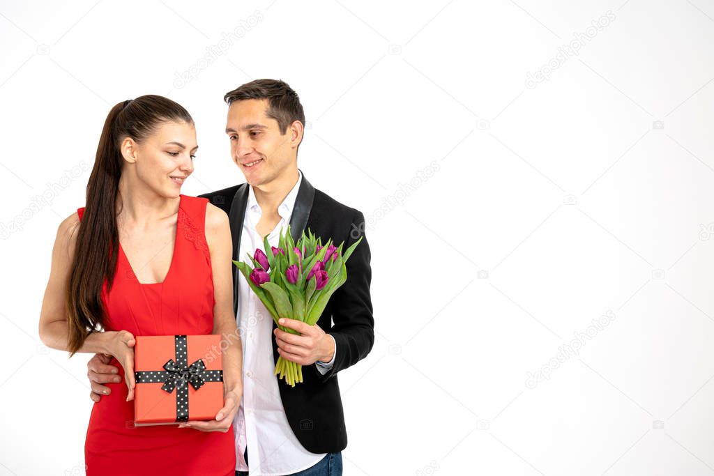 Beautiful romantic couple isolated on white background. Attractive young woman in red dress and handsome man in white shirt are standing with gift box and purple tulips in hands. Happy Saint Valentine