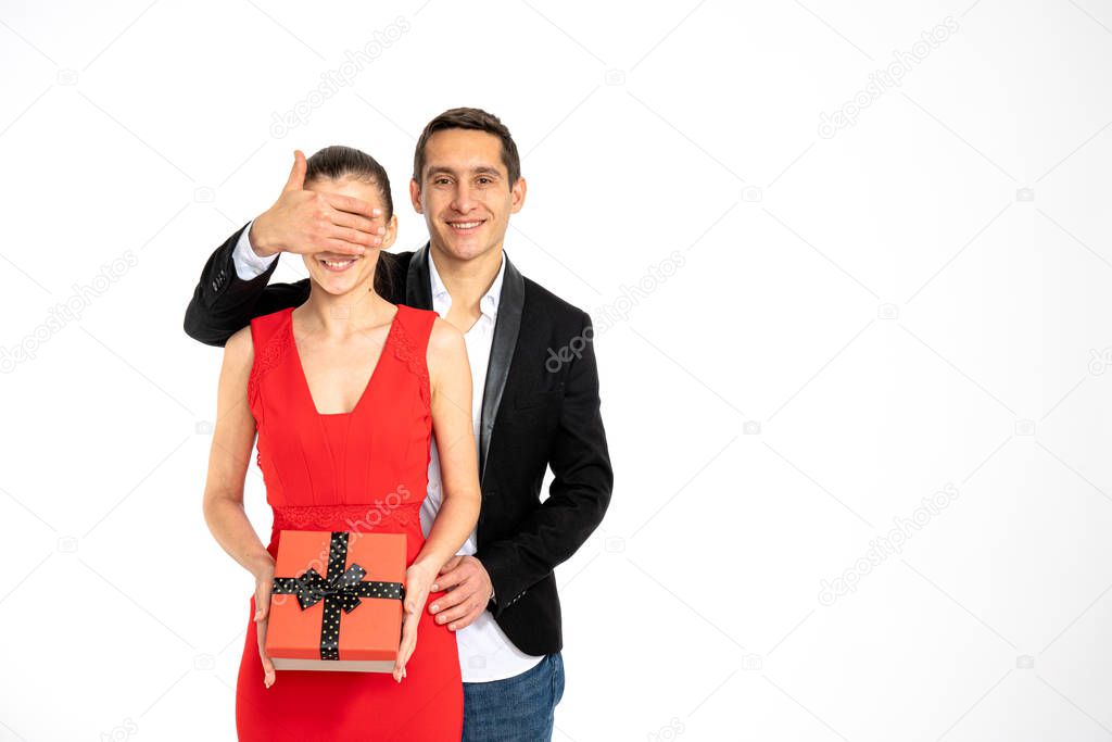 Handsome happy man surprising girlfriend with a gift on white background, elegant couple, male in black blazer covered with his hands his girlfriends eyes smiling and looking at the camera, copyspace