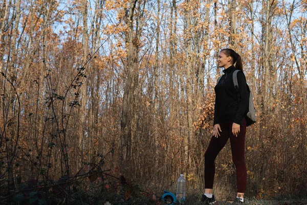 Atractiva chica delgada morena en ropa deportiva oscura, con mochila entre el bosque y mirando al lado izquierdo — Foto de Stock