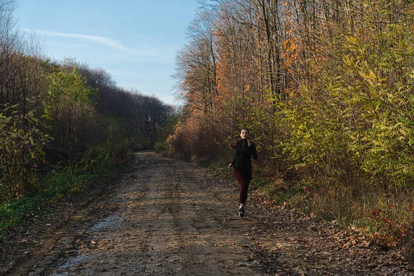 Atractiva chica morena en ropa deportiva oscura corriendo por el camino de tierra entre el bosque — Foto de Stock