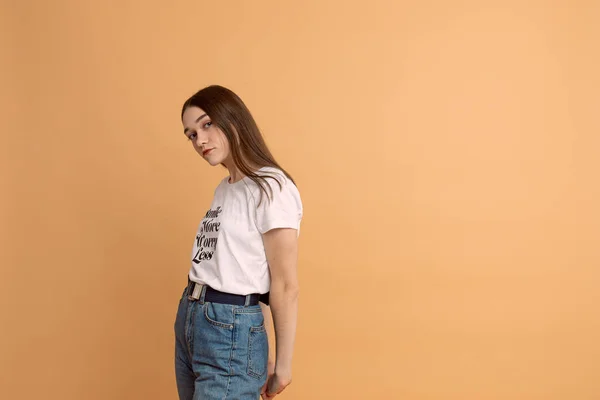 Calmy vážná brunetka dívka v bílém tričku a modré džíny drží ruce za zády a dívá se na fotoaparát izolované přes oranžové pozadí s copyspace — Stock fotografie