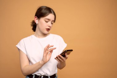 Güzel kız öğrenci cep telefonunu elinde tutuyor, kablosuz kulaklık takıyor, kopyalanmış turuncu arka plan üzerinden izole edilmiş.