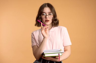 Başarılı kadın yazar ya da gazetecinin görüntüsü, günlüğe not alıyor, kalemi ağzının yanında tutuyor, yuvarlak gözlük takıyor, koyu renk saçlı, turuncu stüdyo duvarında izole edilmiş..