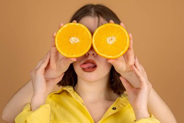 Oyuncu genç kadın dilini gösteriyor, iki çift el turuncu duvar arkasında iki dilim portakalı yüzüne tutuyor.