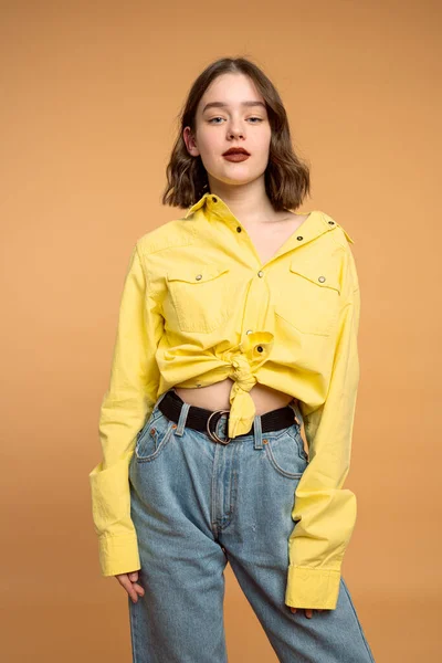 Elegante chica joven con el pelo rizado morena vestida con camisa amarilla mirando a la cámara, aislado sobre fondo naranja —  Fotos de Stock