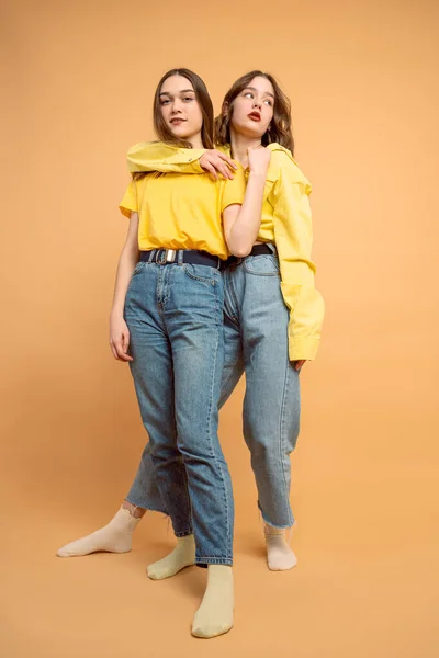 Volledige lengte van twee schattige zelfverzekerde jonge vrouwen in gele shirts knuffelen over oranje achtergrond — Stockfoto