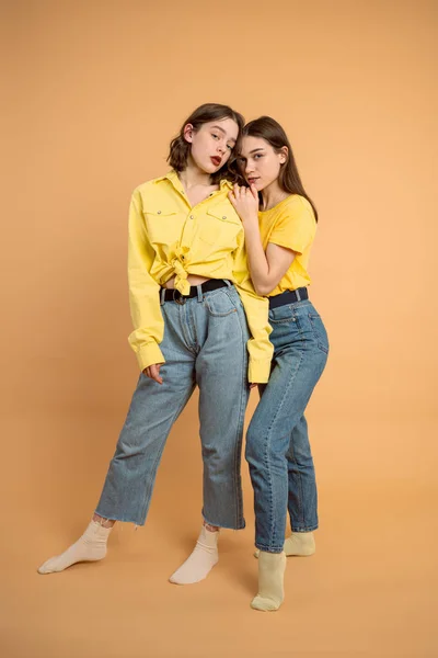 Stijlvolle vrouwen in blauwe jeans en gele shirts die samen staan en naar de camera kijken, geïsoleerd over oranje achtergrond — Stockfoto