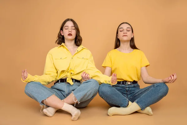 Dos chicas europeas en ropa casual sentadas en el suelo en posición de loto y meditando con los ojos cerrados aislados sobre fondo naranja —  Fotos de Stock