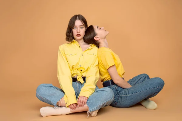 Chicas jóvenes cansadas en ropa casual sentadas en el suelo, apoyadas una sobre la otra, aisladas sobre fondo naranja —  Fotos de Stock