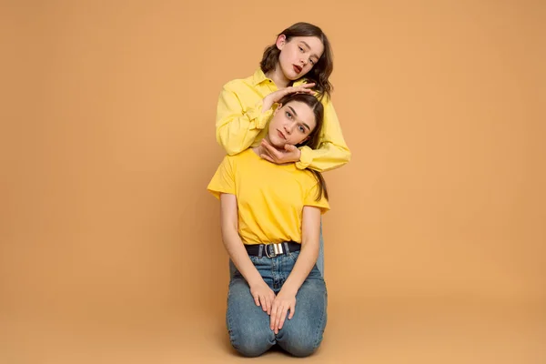 Två attraktiva brunett flickor i gula skjortor sitter på marken en efter en, lockigt hona håller händerna runt ansiktet på sin flickvän, isolerade över orange bakgrund — Stockfoto