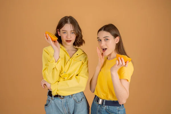 Vackra flickor i gula skjortor som står tillsammans mot orange bakgrund och håller skivade orange i händerna, tittar på kameran — Stockfoto