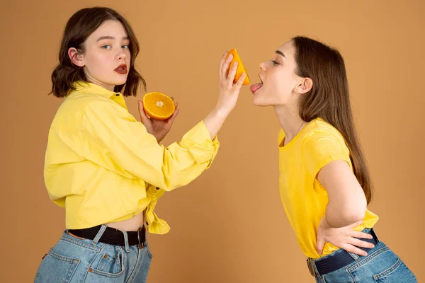 Självsäker flicka i gul skjorta matar sin flickvän med skivad orange och tittar på kameran, isolerade över orange bakgrund — Stockfoto