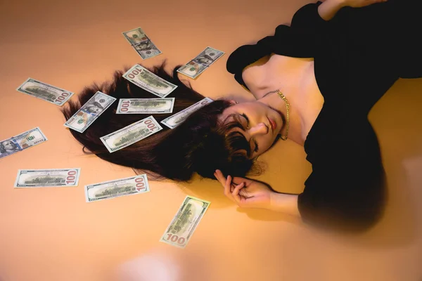 Woman with lot of money. Millionaire woman with brunette loose hair dressed in elegant black dress. Rich sexy woman lies on money. Sexy woman lying in dollar bills