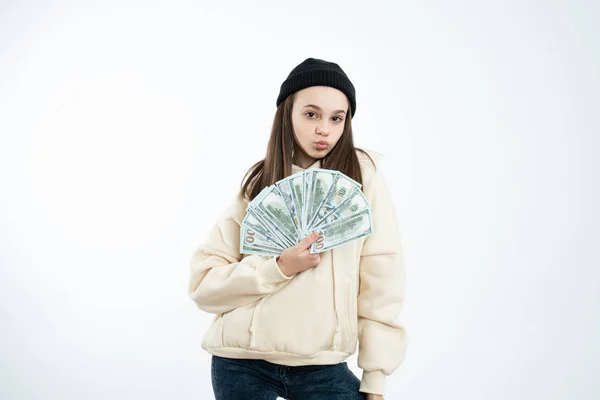 Attractive stylish girl in black hat and beige hoodie holds in her hands dollar banknotes and looking at the camera — Stock Photo, Image