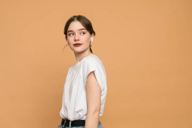 Closeup of a young brunette woman in wireless earphones dressed in white t-shirt, student listening to the music and looking away isolated over orange background with copyspace