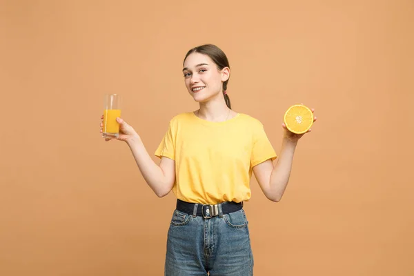 Joyeux belle fille brune en chemise jaune tient l'orange tranchée dans la main droite et un verre de jus dans la gauche, souriant et regardant la caméra — Photo