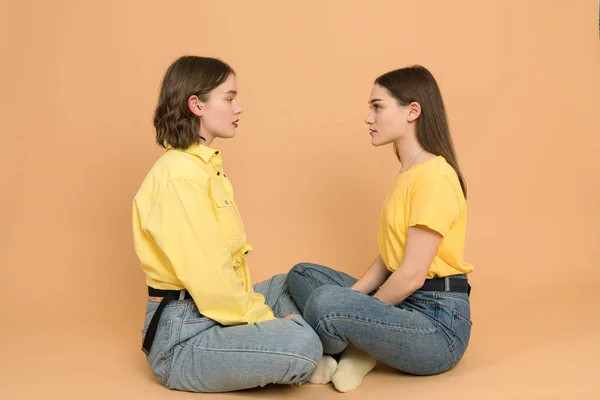 Two attractive girls in blue jeans and yellow shirts sittig against each other and looking into each others eyes, isolated on orange background — Stockfoto