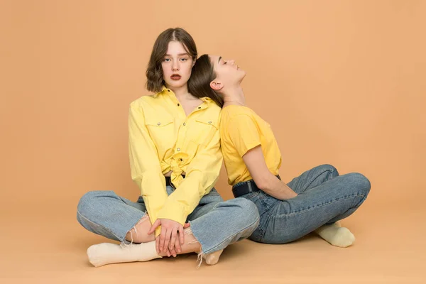 Two young sisters relaxing, woman in casual clothes leaned on her sister in yellow shirt that looking at the camera — Stockfoto