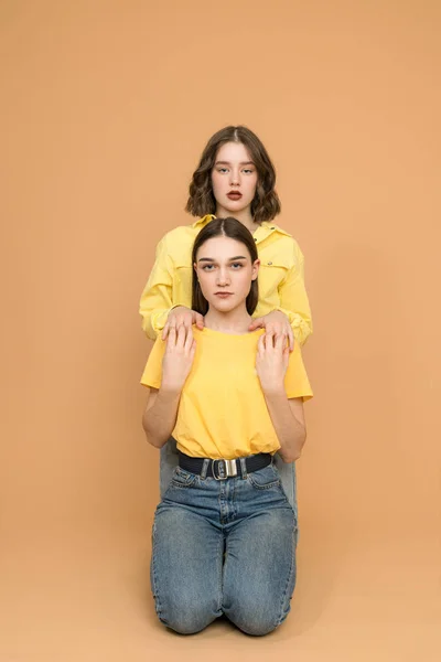 Two brunette sisters sitting on the ground one by one and looking at the camera, isolated over background — Zdjęcie stockowe