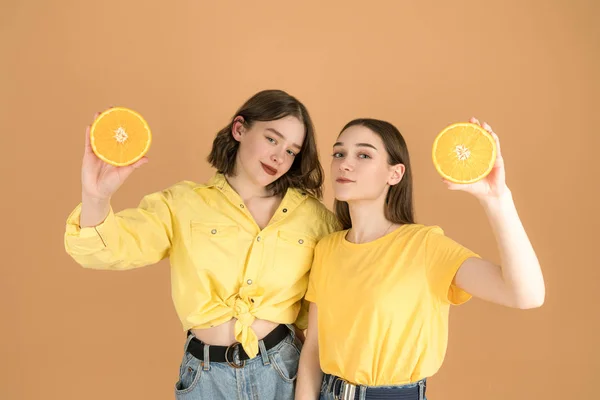 Two beautiful lovly girls in yellow shirts holding sliced orange in their hands and looking at the camera — Zdjęcie stockowe