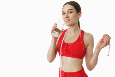 Attractive female in red sportswear drink water, looking at the camera and holds tape measure in her hand, isolated over white background