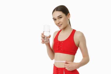 Smiling pleased female in red sportswear holds in her hand glass of water and measuring herself