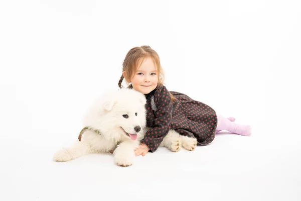 Little kid girl in dress looking at the camera, child hugging her samoyed puppy while liying on the ground — 스톡 사진