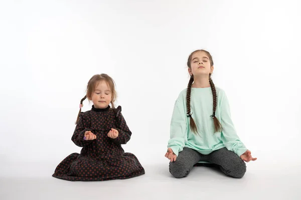 Two little girls in casual clothes sitting on the floor in lotus position with closed eyes and meditating, isolated over white background — Stockfoto