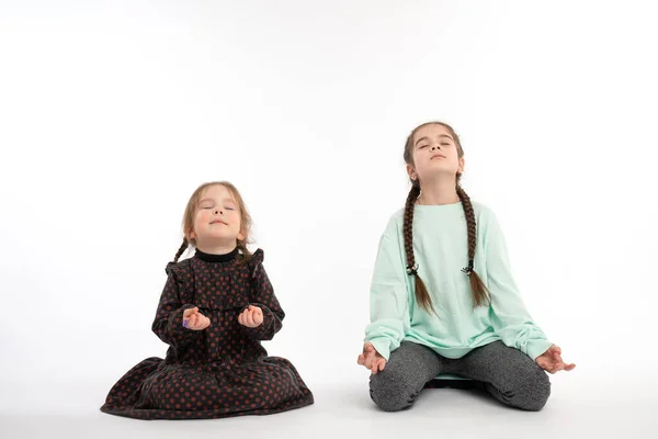 Young girl with pigtails, dressed in blue hoodie teaching her little sister in dress how to meditating, practicing yoga together — Stockfoto