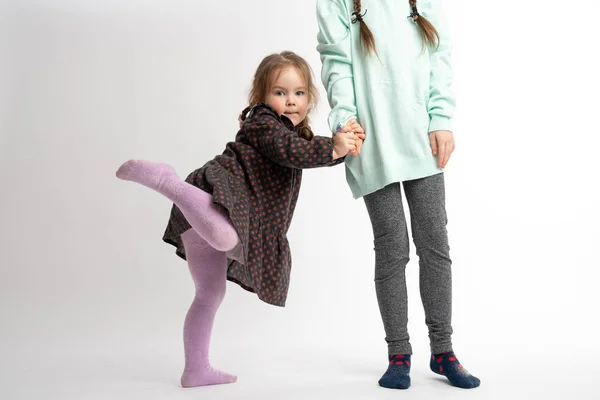 Funny little child girl in dress holds sisters hand and raises her leg, looking at the camera, isolated over white background — ストック写真