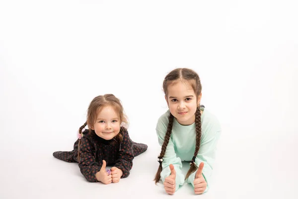 Dos preciosas hermanas con coletas tumbadas en el suelo haciendo un gesto positivo con la mano, con los pulgares hacia arriba y mirando a la cámara aislada sobre fondo blanco —  Fotos de Stock