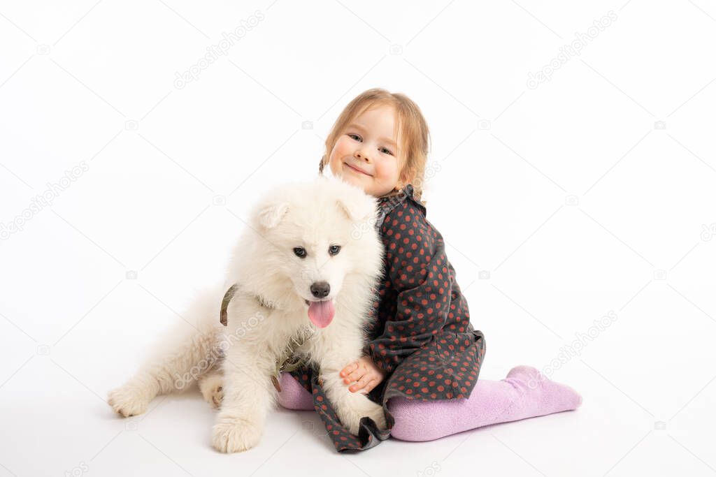 Cute pretty little girl in dress sitting on the ground with white samoyed puppy and looking at the camera