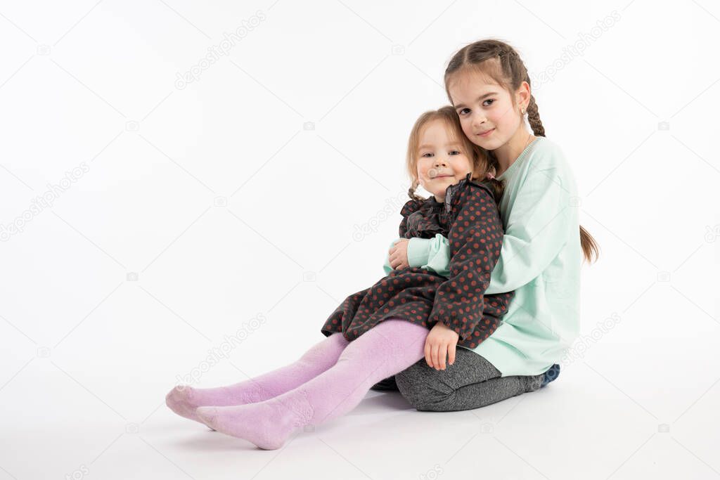 Confident attractive young girl with pigtail, dressed in casual clothes holds her little redhead sister in her arms and looking at the camera, isolated over white background