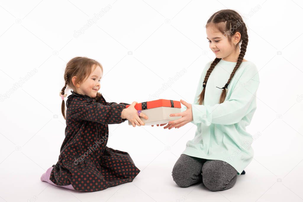Two sisters, little girls with pigtails sitting on the floor, holding present box in their hands and looking at it, isolated over white background