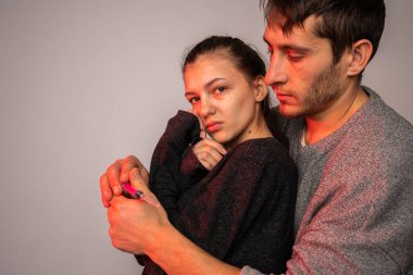 Eski giysiler içinde sakallı bir adam mutsuz karısına sarılıyor ve elinde şırıngayı tutuyor.