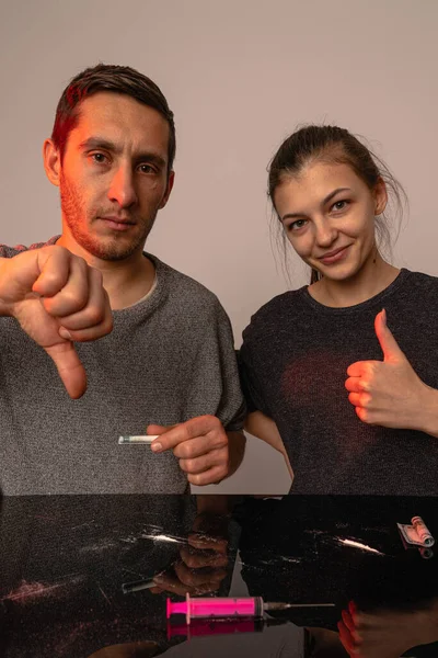 Unhappy man showing thumb down and looking at the camera, brunette female showing thumb up and lokking at the camera, both sitting by the black table with syringe and cocain line