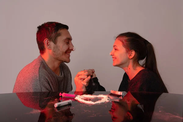 Feliz pareja de drogadictos con polvo blanco en la nariz tomados de la mano y mirándose el uno al otro, hombre y mujer sentados detrás de la mesa con línea de heroína, siringe y billetes — Foto de Stock