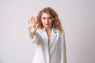 Beyaz ceketli, kırmızı dudaklı, ciddi kıvırcık kız. Avuç içi açık, dur işareti veriyor. Gri arkaplanda izole edilmiş kameraya bakıyor.